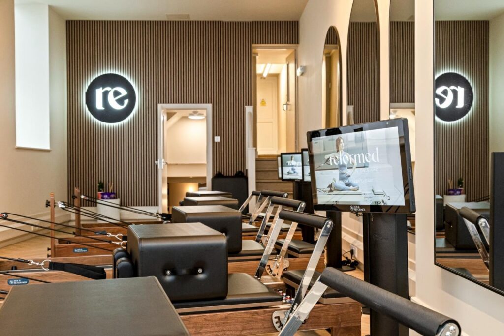 Studio perspective with illuminated ‘re’ logo wall, reformers and arched mirrors.